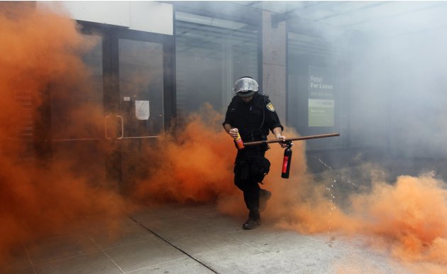 A police officer in riot gear emerges from the colored smoke in front of destroyed storefronts during May Day demonstrations