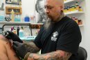 In this Thursday, June 27, 2013, photo, former Fort Knox soldier "Baldy" Carder inks a customer's arm at his Radcliff, Ky., tattoo parlor. Businesses like Carder's could be affected by the recent move by the Army to eliminate a brigade of about 3,300 soldiers at the famed Army post. (AP Photo/Dylan Lovan)