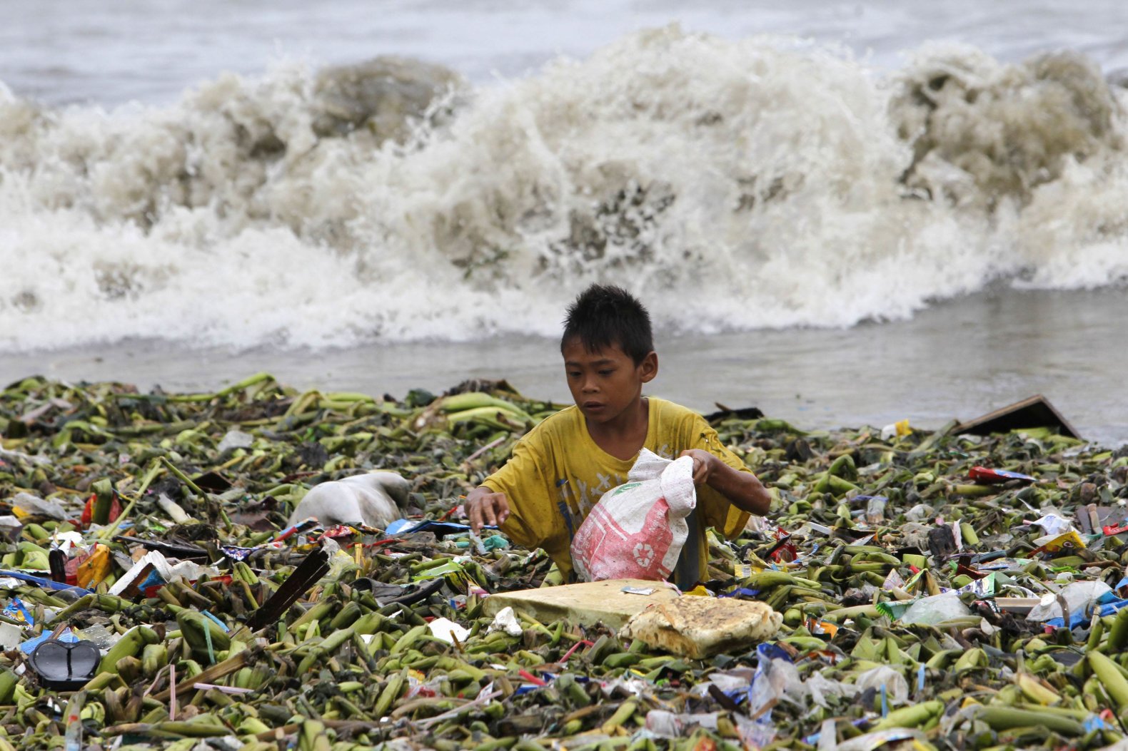 A boy sifts through floating garbage as he collects recyclable items to sell while strong waves crash along the shores of Manila Bay, near a slum area in Manila